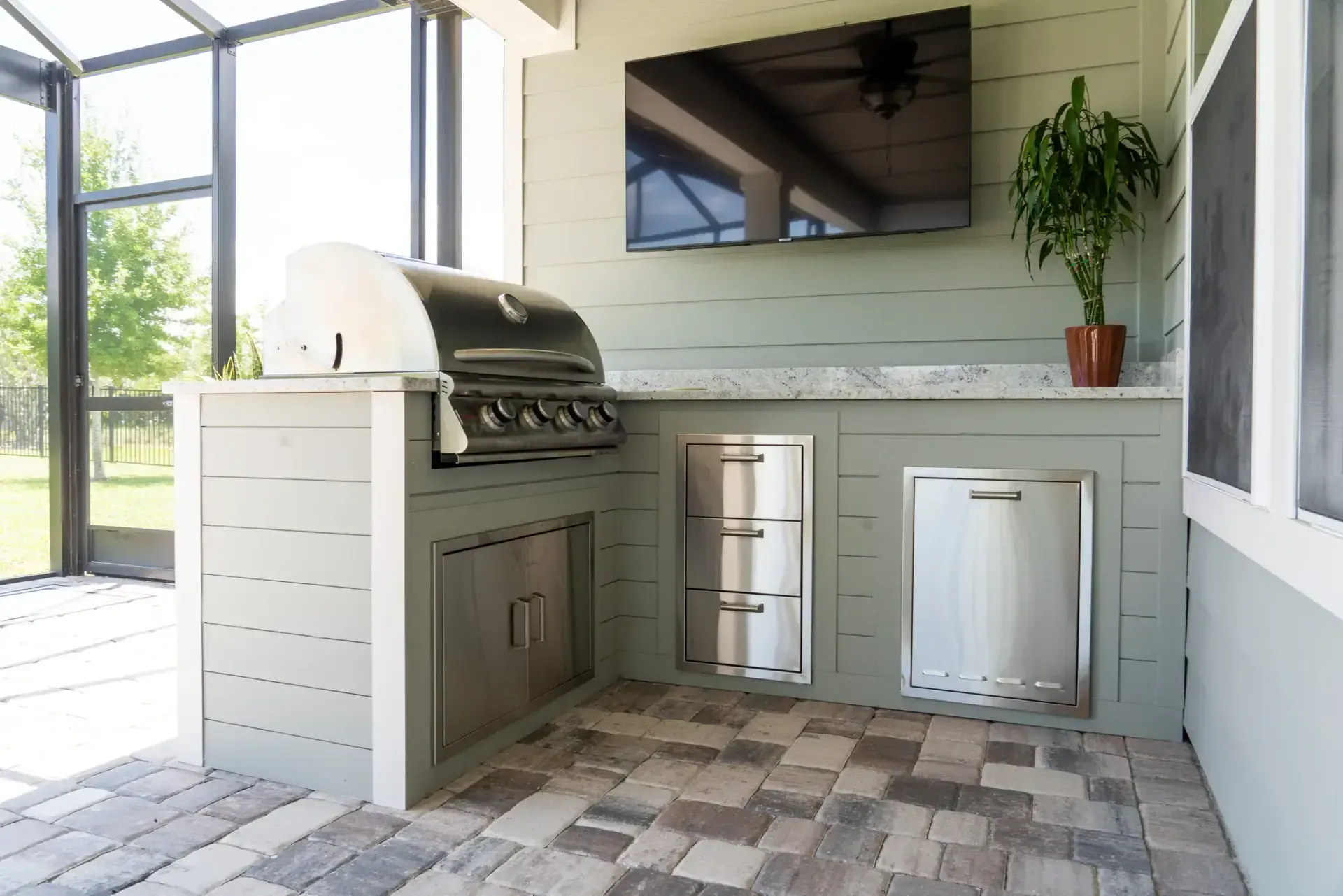 A shiplap-faced outdoor kitchen with grill and television over a paver floor